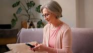 Mature old 60s woman, older middle aged female customer holding smartphone using mobile app, texting message, search ecommerce offers on cell phone technology device sitting on couch at home