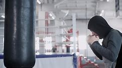 Boxer in hoodie is boxing hitting punches on punching bag in empty gym, side view. Young fighter is training preparing to competitions. Workout, martial art, training, sport, boxing, fighting concept.