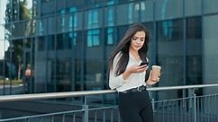 Elegantly dressed beautiful attractive woman working in a corporation, a company, came out in front of a glass modern building for break, holds cup of takeaway coffee and phone, reads news