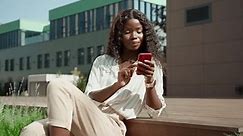 Young African happy woman customer holding cell phone using mobile apps for ecommerce shopping, online dating applications, communicating online on cellphone tech device sitting outside.