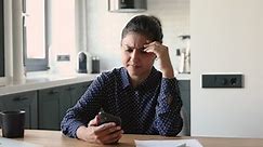 Indian ethnicity woman sit at desk holding smart phone feels unhappy negative emotions upset by receiving terrible news by sms. Device damaged, lost internet connection, need gadget fix repair concept