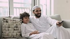 Happy Muslim father and cute little 5s son in traditional clothing sit on couch relaxing watching cartoons on on-line streaming TV services. Smiling Islamic dad and child enjoy family weekend at home