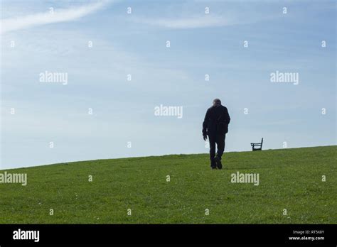 An Older Male Figure Walks Across Some Open Grass Towards A Park Bench