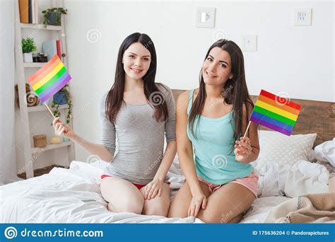 Lesbian Couple In Bedroom At Home Sitting Holding Lgbt Color Flags