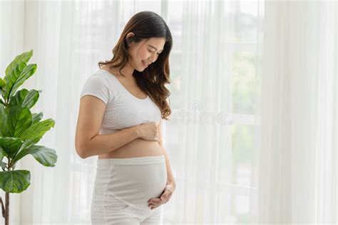 Happy Pregnant Woman Standing In Front Of Windows And Stroking Her Big