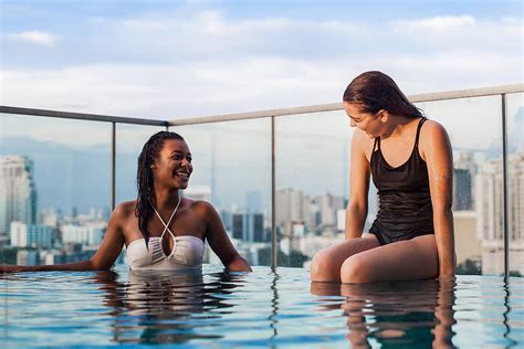 Two Girlfriends Relaxing At Swimming Pool With City Skyline Behind Them