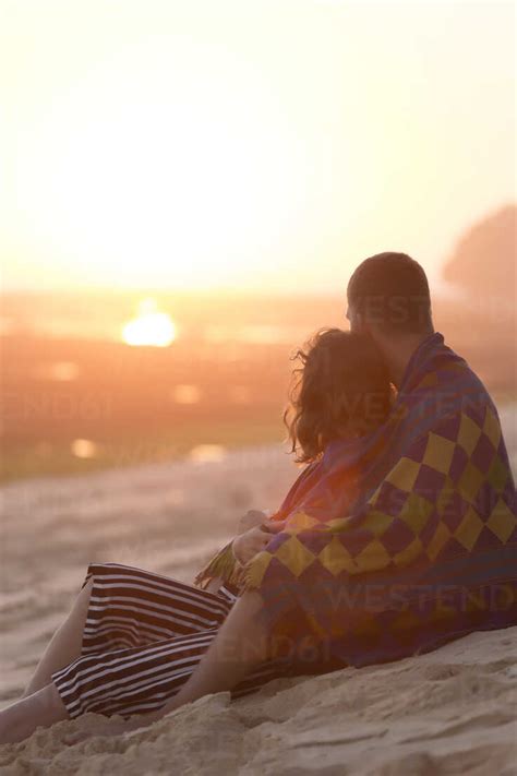 Thoughtful Young Couple Wrapped In Blanket While Sitting At Beach
