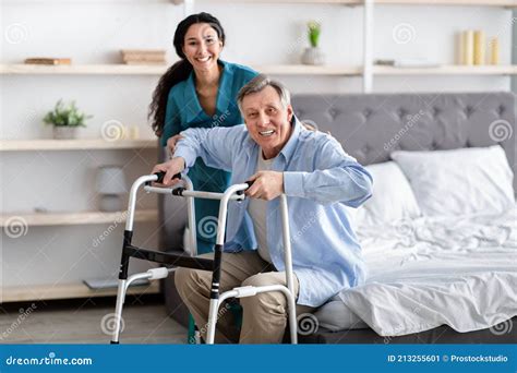 female nurse helping elderly male with walking frame stand up from bed