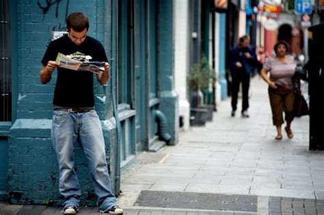 A Man Standing On The Sidewalk Reading A Newspaper