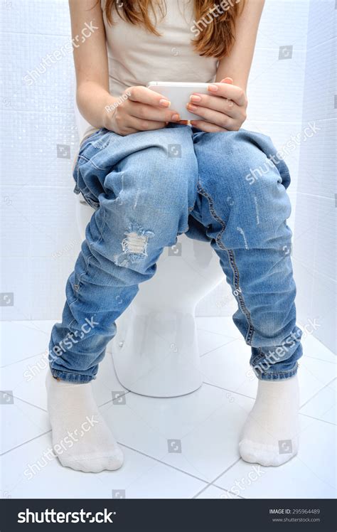 Young Woman Sitting On Bathroom Or Wc Toilet Bowl Using Phone In Hands