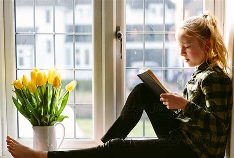 pre teen girl sitting in a sunny window reading by stocksy