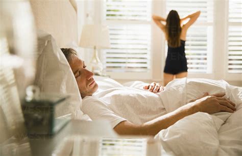 wife stretching at morning window behind husband sleeping in bed stock