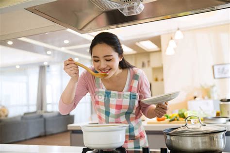 Smiling Young Woman Cooking And Tasting Dish In Kitchen — Homey Asian