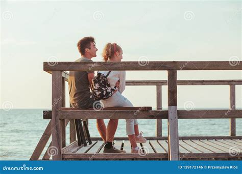 Happy Smiling Couple Of Young Woman And Man Sitting On A Pier Over The