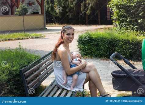 Smiling Happy Mother Sitting On Park Bench And Breastfeeding Hungry