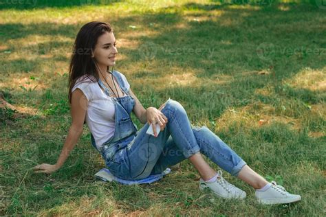 Slender Brunette Teen In Jeans Overalls Lying On The Grass In The Park