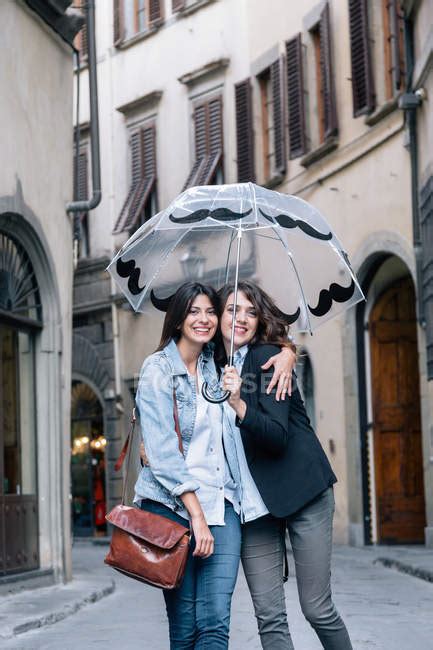 lesbian couple standing together in street holding umbrella looking at