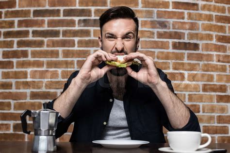 Handsome Bearded Young Man Eating Sandwich And Drinking Coffee At