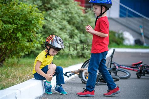 小男孩从自行车上摔了下来。不高兴的男孩坐在柏油路上看着他的膝盖。他的朋友扶他站起来，又骑了起来照片摄影图片_ID:353434189-Veer图库