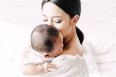 母亲抱着婴儿在她的怀里, 亲吻在一个白色的卧室。对家庭概念的爱照片-正版商用图片0s5mrb-摄图新视界