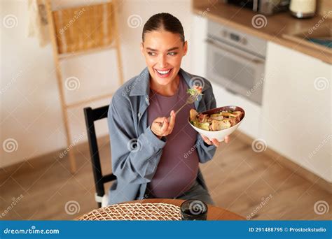 Satisfied Pregnant Woman Eating Caesar Salad, Sitting at Table in Kitchen, Holding Bowl, Looking ...