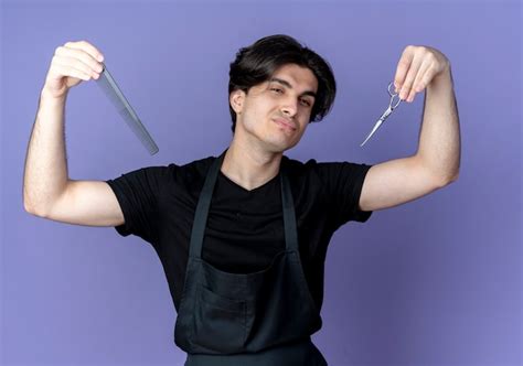 Confident young handsome male barber in uniform raising scissors with ...