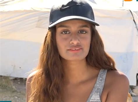 Shamima Begum leaning against a door frame. She has long dyed brown hair and is wearing sunglasses with a light grey top underneath a sweatshirt jacket