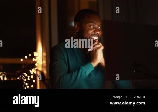 A man with dark skin sits at a desk, smiling while looking at a computer screen. The warm light creates a cozy atmosphere at night. He appears focused and content with his work Stock Video Footage - Alamy