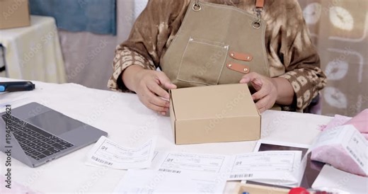 Senior asian artisan attaches shipping label on carton, aligns barcode sticker with invoice, checks online order record on laptop, prepares parcel packaging for delivery from home studio workspace.