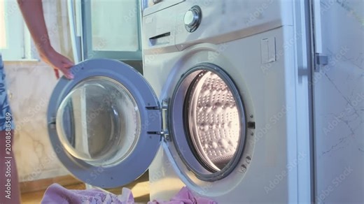 Woman placing dirty clothes into front loading washing machine at home. Domestic routine, laundry process, preparing garments for wash, everyday household chores and cleaning lifestyle.