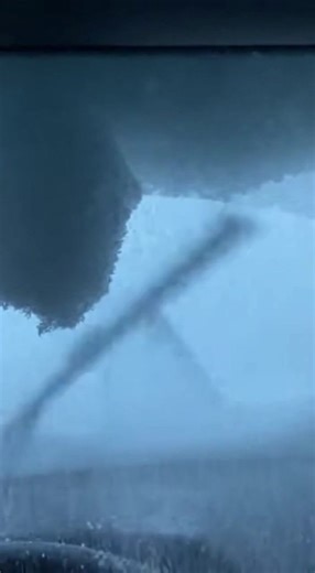 Avalanche of Snow Collapses Into Highway Cut, Burying Cars Mid-Drive Mountain Pass — A routine winter drive through a mountain highway cut turned into a whiteout nightmare when a massive wall of snow collapsed from above, spilling into the roadway and rapidly covering vehicles trapped below. The video shows traffic creeping through a narrow corridor carved into solid rock, with towering snowbanks rising on both sides. Ahead, an overhanging shelf of snow hangs ominously across the gap like a froz