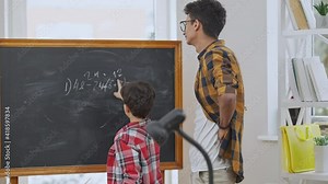 Young smiling Middle Eastern teacher in eyeglasses listening to pupil explaining math problem solution written on chalkboard. Positive tutor teaching schoolboy in classroom. Slow motion.