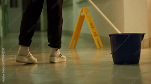 Close-up of a janitor mopping a tiled hallway floor with a bucket and caution sign visible, emphasizing workplace hygiene and safety. Concept of professional cleaning and maintenance.