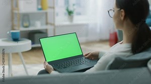 Young Woman at Home Sitting on a Couch Works on a Laptop Computer with Green Mock-up Screen. Girl Using Computer, Browsing through Internet, Watching Content, Chatting in Social Networks with Friends.
