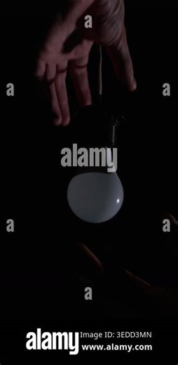 A close-up view of a person hand carefully screwing a white LED lightbulb into a hanging socket surrounded by complete pitch darkness. The lamp subtly flickers as electrical contact is made Stock Video Footage - Alamy