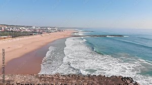 Beauty of an ocean coastline from a drone's perspective. The camera ascends, revealing teal waves lapping against the sandy beach, a scenic promenade, wavebreakers, and a charming town in the distance