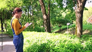 Young beautiful asian woman natural botany student, plant researcher walking, studying plants and herbs, medicine in the garden writing down the names of plants on a clipboard with interest.