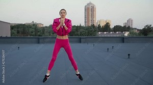 charming sportswoman holding hands on hips, jumping and squatting on rooftop, looking at camera