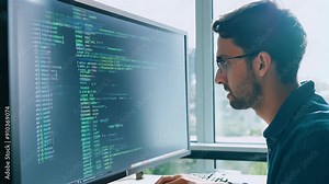 A programmer wearing glasses works at a computer with code displayed on the screen. Concept: programmer's work, software development, writing code,