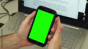 Anonymous man in front of his computer makes gestures with his hands holding a mobile phone or cellphone with a greenscreen or chroma key screen seems angry or annoyed with what he sees.