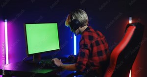 Rear of the Caucasian blonde female computer games gamer in the headphones playing at the big chroma key screen in the dark neon gaming room. Back view. Alpha channel. Green screen.