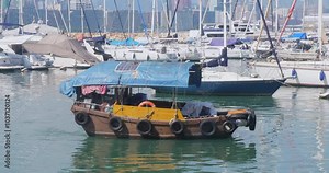small boat with a blue canopy and solar panel, lined with blacktires floating in a marina, among other boats and yachts docked in the background