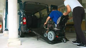 Man on wheelchair using accessible vehicle with ramp for transportation with driver helping.