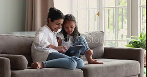 Indian woman and 6s daughter use digital tablet, choose goods for kids online, buying in e-commerce retail services sit on sofa at home. Young generation use modern tech for development, fun concept