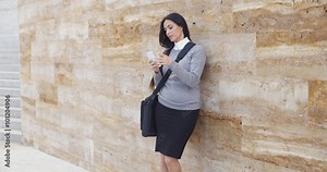 Serious young business woman in gray sweater and black skirt with briefcase checking email on her phone while leaning against a large marble wall