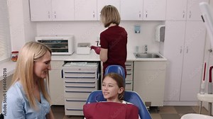 Dentist talking, Dental background, Clinic environment. Dentist in blue scrubs talking to young girl in dental chair, with dental assistant in red scrubs preparing tools in background.