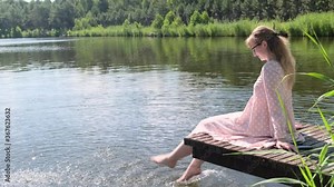 Woman on the lake. Young woman in pink dress sitting on a wooden platform on the lake and dip her feet in water, creating splashes, slow motion