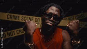 Young Black rapper wearing sunglasses and gold accessories performing hip-hop song expressively in front of crime scene tape in flashing police lights at night