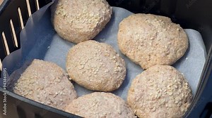 Whole wheat dough buns sprinkled with sesame seeds are arranged on parchment paper in an air fryer basket, ready for baking. The close-up shot highlights the texture and preparation stage of the buns