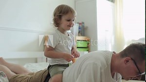 A little blond boy with big brown eyes sits on his father's back imagining that this is a horse while his father does an exercise and push-up off the floor. Close-up